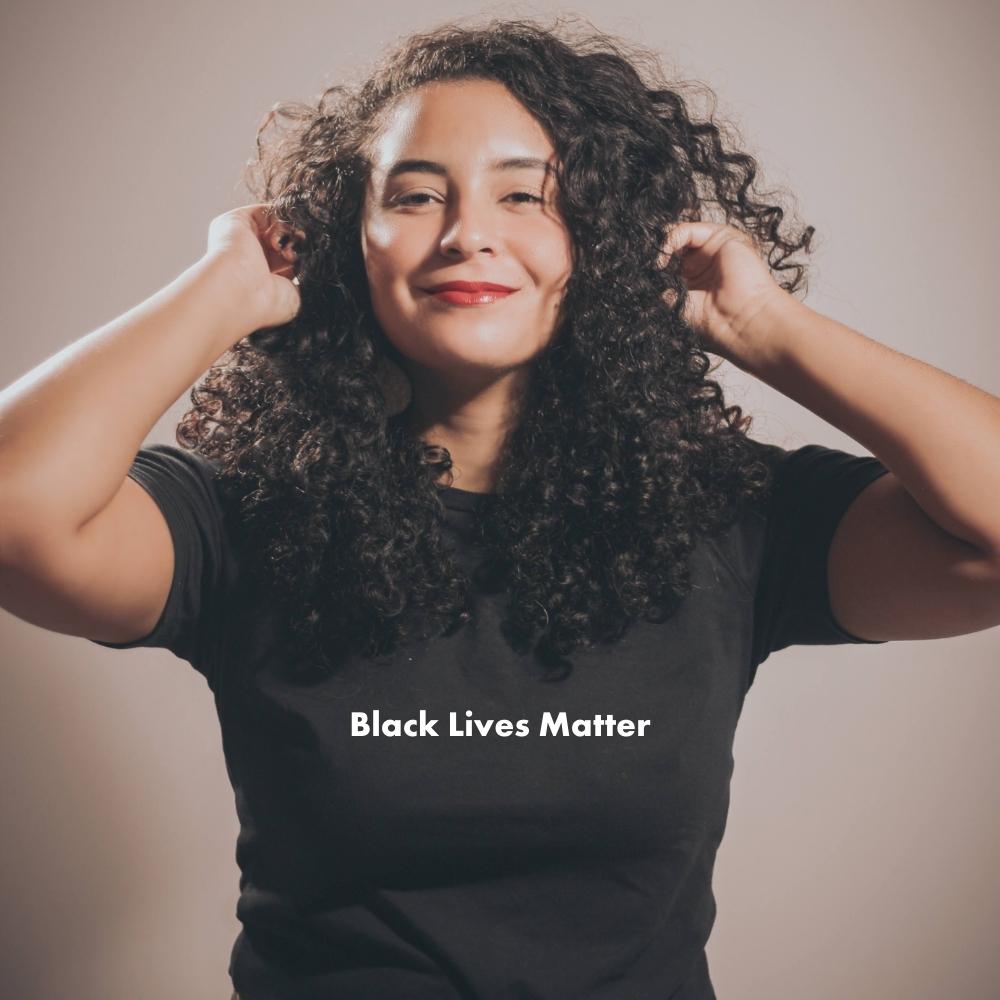 A woman with long, curly hair is posing for a photo with both of her hands on either side of her head, interlocking with her curls. She is wearing a black t-shirt that reads "Black lives matter" on it in white writing.