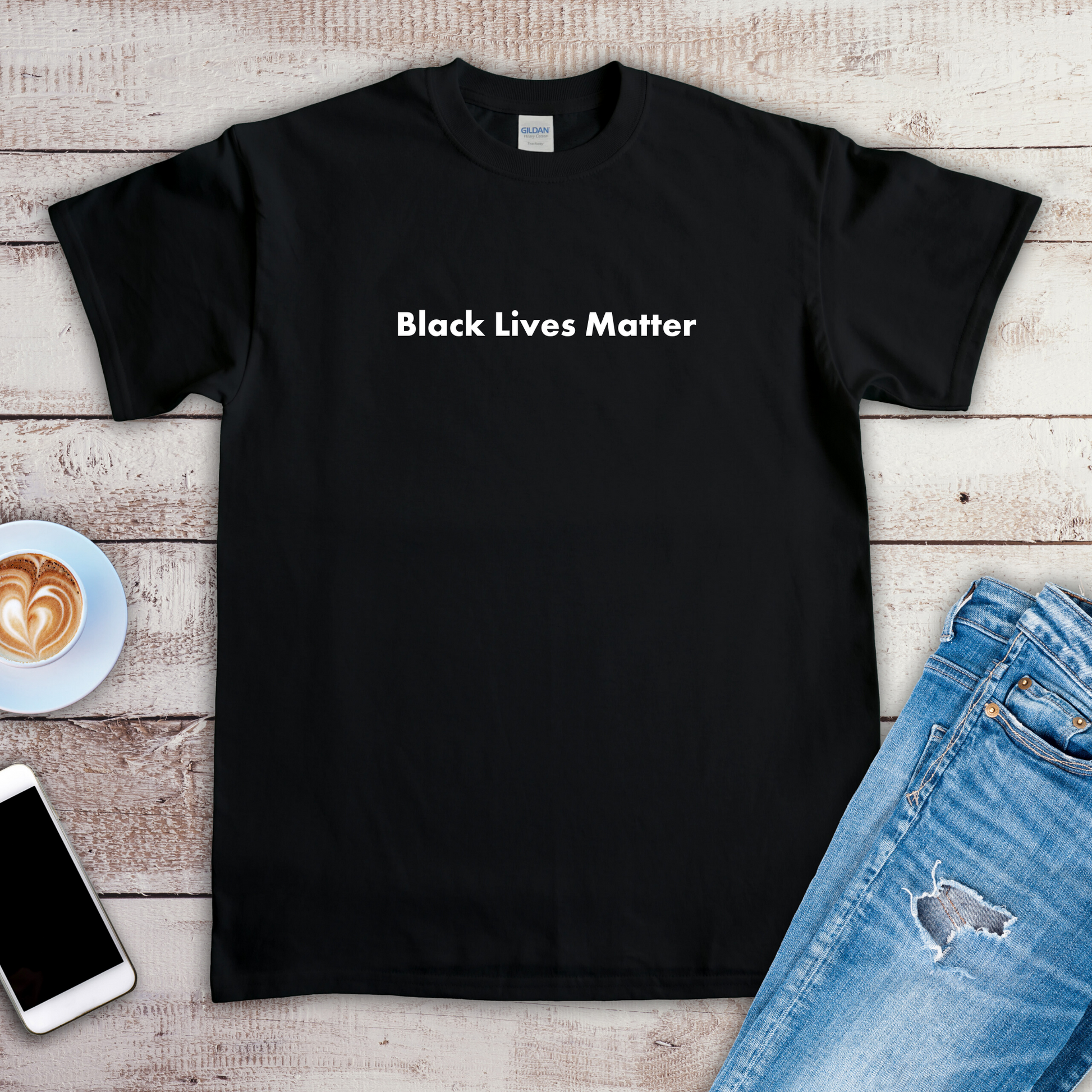 A black t-shirt is laying on a wooden floor. The t-shirt has a statement on it that says "Black lives matter". There are a pair of jeans, a phone, and a cup of coffee next to the t-shirt.