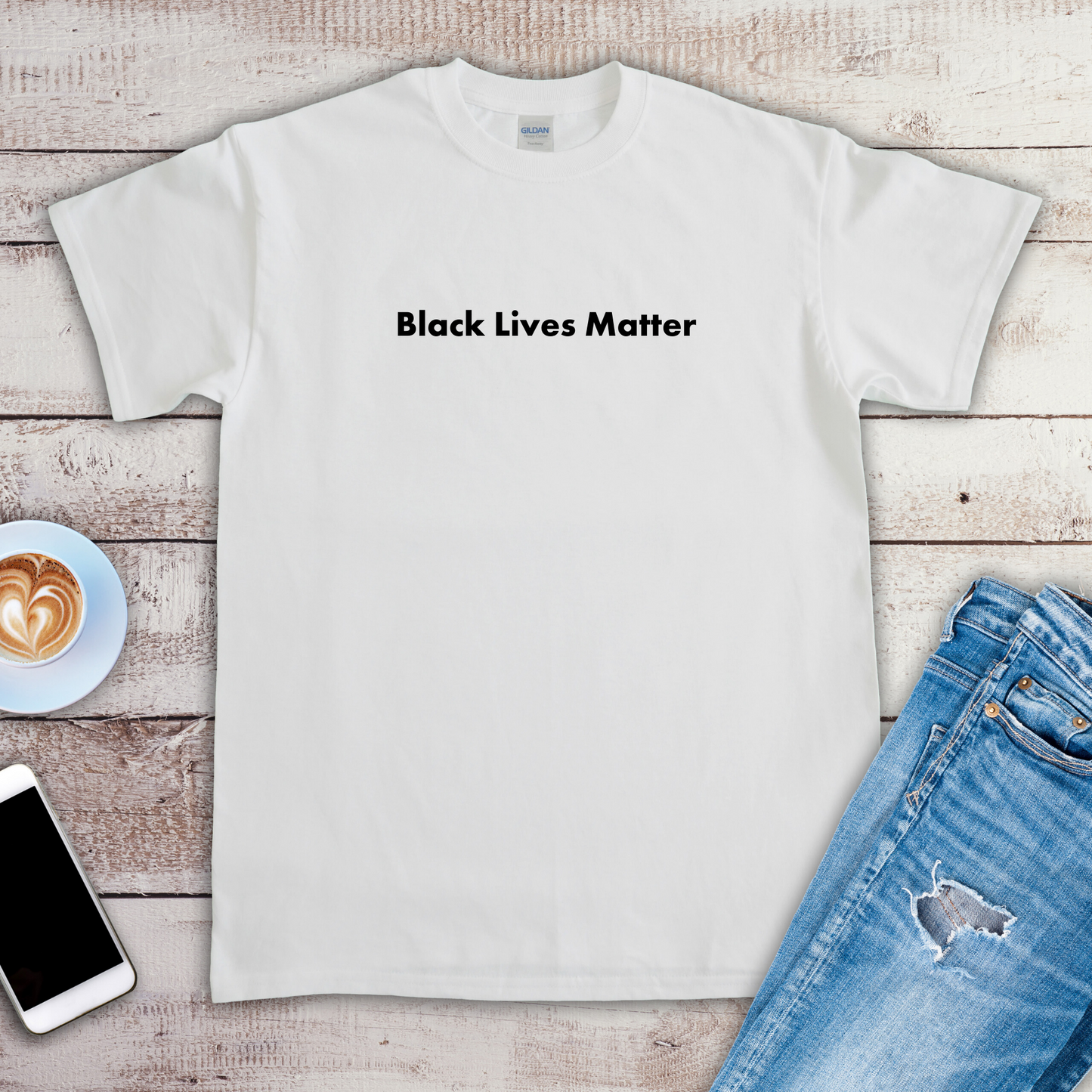 A white t-shirt is laying on a wooden floor. The t-shirt has a statement on it that says "Black lives matter". There are a pair of jeans, a phone, and a cup of coffee next to the t-shirt.