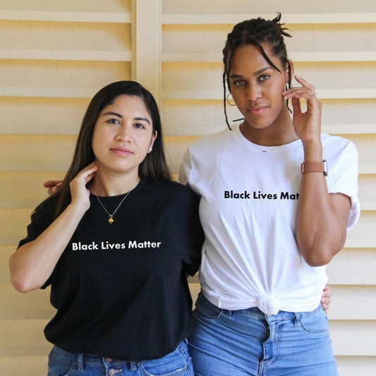 Two women standing side by side posing for a picture. The shorter woman is wearing a black t-shirt that says "black lives matter" on it in white. The taller Black woman is wearing a white t-shirt that says "black lives matter" on it in black.