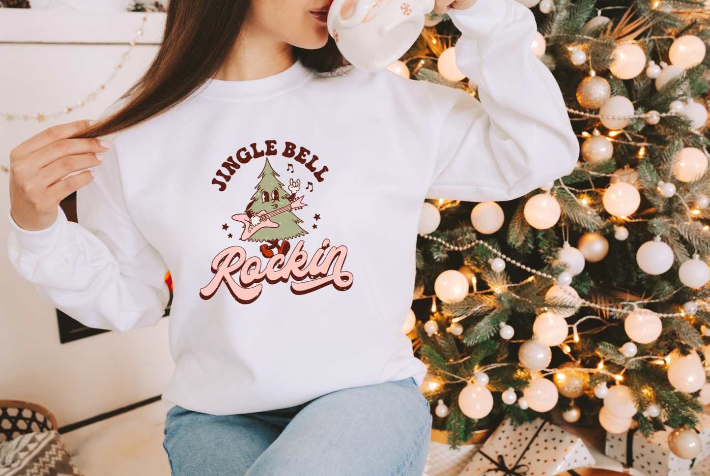 A woman standing in front of a Christmas tree drinking out of a coffee mug and wearing a shirt that displays a christmas tree character playing a guitar.