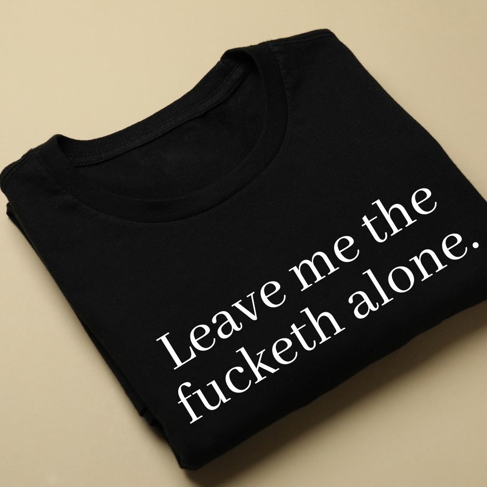 A black crewneck sweatshirt is folded up and laying on a light yellow background. It has a funny, sarcastic saying on it "leave me the fucketh alone" in white writing.