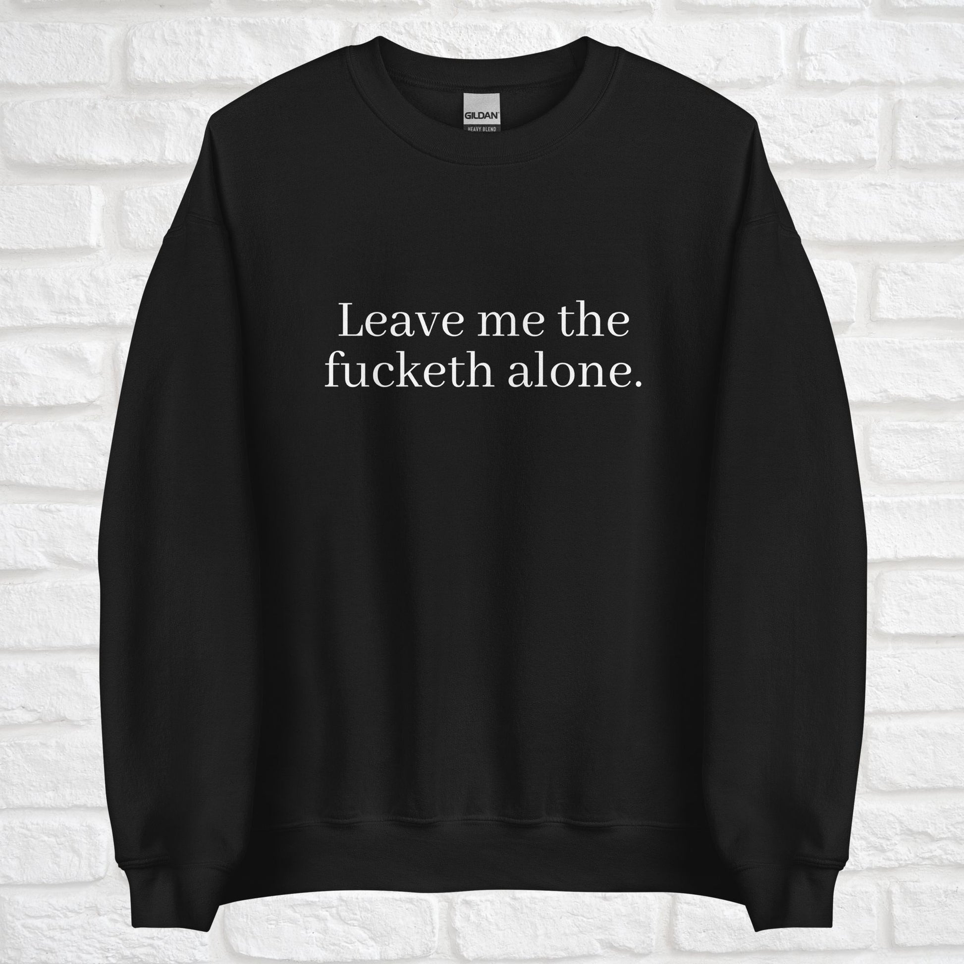 A black crewneck sweatshirt has a funny saying on it in white that says "leave me the fucketh alone". The sweatshirt is tapered at the bottom of the shirt and sleeves. White bricks are in the background.