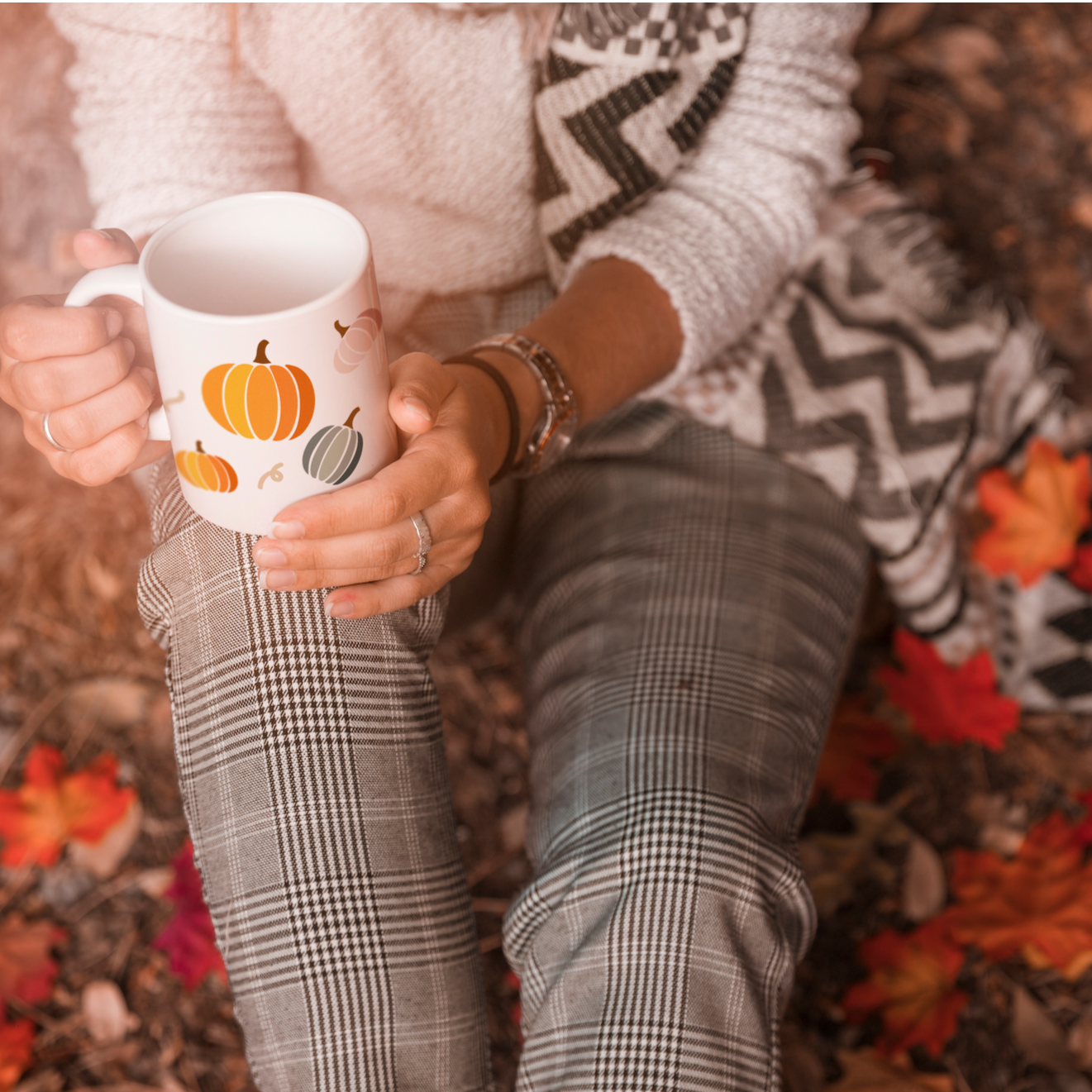 woman sitting on the ground in a forest on red and orange autumn leaves holding a white mug with an image of a colourful orange and green pumpkin patch