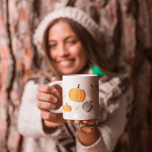 woman with a knitted hat standing in front of a tree holding a white mug with an image of a colourful orange and green pumpkin patch